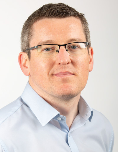 Professional headshot of a man with short brown hair and glasses, wearing a light blue button-down shirt.